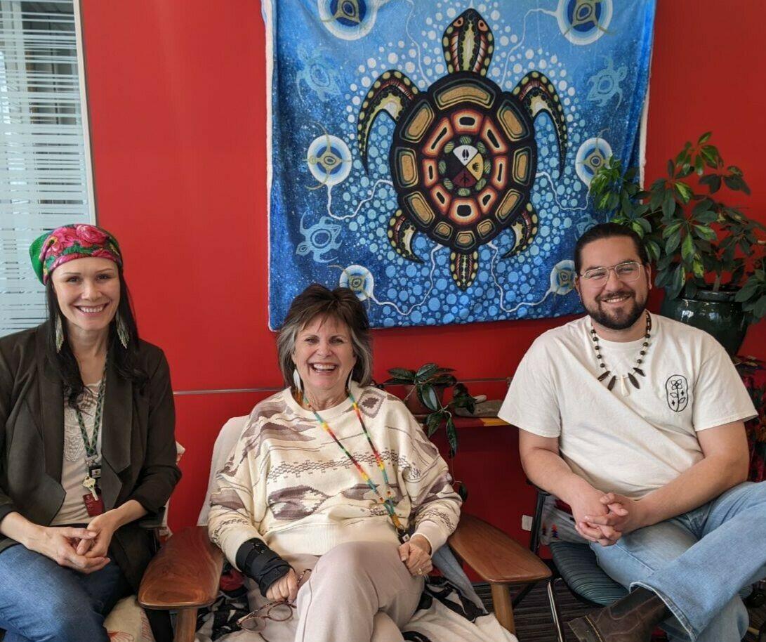 three people sit against a wall with Indigenous artwork behind them
