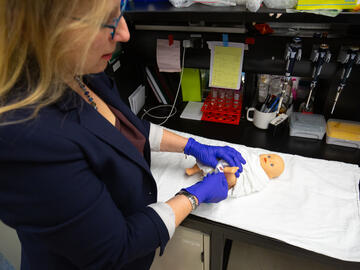 A woman wearing gloves holds a 'bloodspot' next to a newborn doll